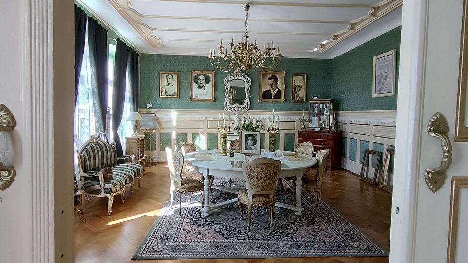 Blick durch eine geöffnete Flügeltür in ein historisch eingerichtetes Salon- oder Speisezimmer mit grüner Tapete, weißer Wandvertäfelung und Stuckdecke samt Kronleuchter. In der Mitte steht ein runder Tisch auf einem gemusterten Teppich, umgeben von Polsterstühlen; an den Wänden hängen gerahmte Porträts, daneben stehen eine Vitrine und ein Sofa vor hohen Fenstern mit dunklen Vorhängen.