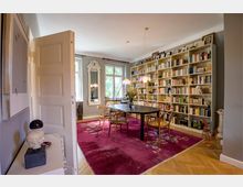 Elegantes Wohnzimmer mit grauen Wänden, großem Bücherregal, ovalem Esstisch und Holzstühlen auf einem blumenverzierten, roten Teppich. Im Hintergrund stehen ein bodenhoher Spiegel und ein Fenster, das Tageslicht in den Raum lässt.