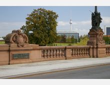 Sandsteinbrücke mit verzierten Balustraden und Skulpturen, im Hintergrund ein moderner Bürokomplex und der Berliner Fernsehturm, umgeben von Bäumen und einer Parklandschaft.