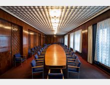 Großer Konferenzraum mit einer zentralen ovalen Holztafel und blauen Stühlen, umgeben von hölzernen Wandverkleidungen mit dekorativen Musterausschnitten. Die Decke ist mit geometrischen weißen Elementen gestaltet, und die rechte Seite ist von hohen Fenstern mit Spitzenvorhängen gesäumt.