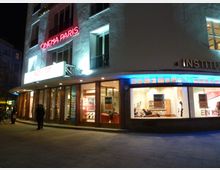 Straßenansicht eines modernen Gebäudes bei Nacht mit dem beleuchteten Schriftzug "Cinema Paris" über einem Eingang. Die Fassade besitzt große Fensterfronten, durch die ein einladender Innenraum mit Sitzbereichen sichtbar ist.