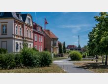 Eine ruhige Wohnstraße mit mehrstöckigen, historischen Häusern in verschiedenen Farben, umgeben von gepflegten Hecken und Bäumen. Im Vordergrund verläuft ein asphaltierter Gehweg, und im Hintergrund sind weitere Wohnhäuser sowie ein klarer, blauer Himmel zu sehen.