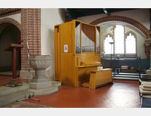 Innenraum einer Kirche mit einer Orgel aus hellem Holz, einem steinernen Taufbecken und einem Rednerpult auf einem mobilen Gestell. Im Hintergrund sind gotische Fenster mit roten Backsteinbögen und Sitzbänke zu sehen.