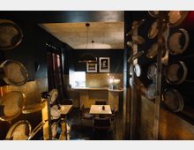 Dunkel beleuchteter Innenraum eines kleinen Restaurants mit modernem Design. Zu sehen sind Tische mit Stühlen, eine Theke im hinteren Bereich, dekorative Metallplatten an den Seitenwänden und Pendelleuchten unter einer dunklen Decke.