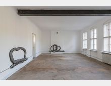 Ein leerer Raum mit weißen Wänden und mehreren hohen Fenstern auf der rechten Seite, die Tageslicht hereinlassen. Sichtbare Kunstinstallationen aus Metallrohren sind an den Wänden befestigt, der Boden ist roh und stellenweise fleckig.