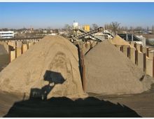Sandlagerplatz auf einer Baustelle mit großen Sandhügeln, umgeben von Metallwänden und industriellen Fördereinrichtungen im Hintergrund; im Vordergrund fällt der Schatten eines Radladers auf den Sand. Im Hintergrund sind Gebäude und Bäume sichtbar, bei klarem, sonnigem Wetter.