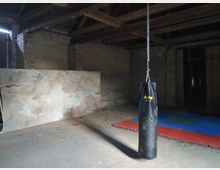 Ein leerer Raum mit einer schwarzen Boxsacktasche, die an einer Kette von der Decke hängt. Im Hintergrund stehen eine große bemalte Tafel mit Wolkenmotiv, freiliegende Ziegelwände und bunte Turnmatten auf dem Boden.