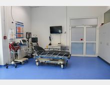 Ein moderner Behandlungsraum in einer Klinik mit blauem Boden, einer medizinischen Liege, Monitoren und Geräten für Überwachungs- und Notfallversorgung. Im Hintergrund befinden sich ein Wandmonitor, ein Schrank und Türen mit Milchglas.