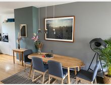 Esszimmer mit Holztisch und vier gepolsterten Stühlen auf einem Teppich. An der grauen Wand hängen Bilder, darüber drei Hängelampen, daneben stehen Pflanzen, eine Stehlampe und ein kleiner Tisch mit Dekoration. Im Hintergrund ist eine weiße Küchenzeile sichtbar.