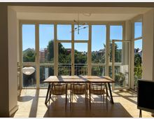 Bright dining area with a wooden table and chairs set in front of large floor-to-ceiling windows and a glass door opening to a small balcony. White-framed windows fill the wall, letting in sunlight and views of trees and nearby rooftops, with a modern ceiling light fixture overhead.