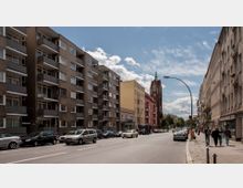 Straßenszene in einer Stadt mit mehrstöckigen Wohngebäuden auf beiden Seiten, geparkten Autos entlang der Straße und Gehwegen. Im Hintergrund steht ein rotes Kirchengebäude mit einem grünen Turm, umgeben von weiteren Gebäuden und bewölktem Himmel.