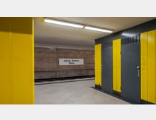 Untergrundbahnhof mit gelben und grauen Wandverkleidungen sowie einer Beschilderung mit der Aufschrift „Jakob-Kaiser-Platz“ auf einer Betonwand. Der Boden ist mit hellgrauen Fliesen gestaltet, und die Beleuchtung erfolgt durch Deckenlampen.