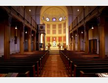 Innenraum einer Kirche mit einem zentralen Mittelgang, flankiert von Holzbankreihen. Im Hintergrund befindet sich ein Altar mit einem Kreuz, flankiert von Blumenarrangements, und darüber Rundfenster mit farbigen Glasornamenten.