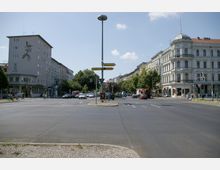 Straßenkreuzung in einer Stadt mit mehrspuriger Fahrbahn, mehreren Verkehrsschildern und Ampeln. Um die Kreuzung herum stehen Wohn- und Geschäftshäuser, darunter ein Eckgebäude mit Kuppel und ein modernes Gebäude mit Wandkunst, sowie Bäume und geparkte Autos.