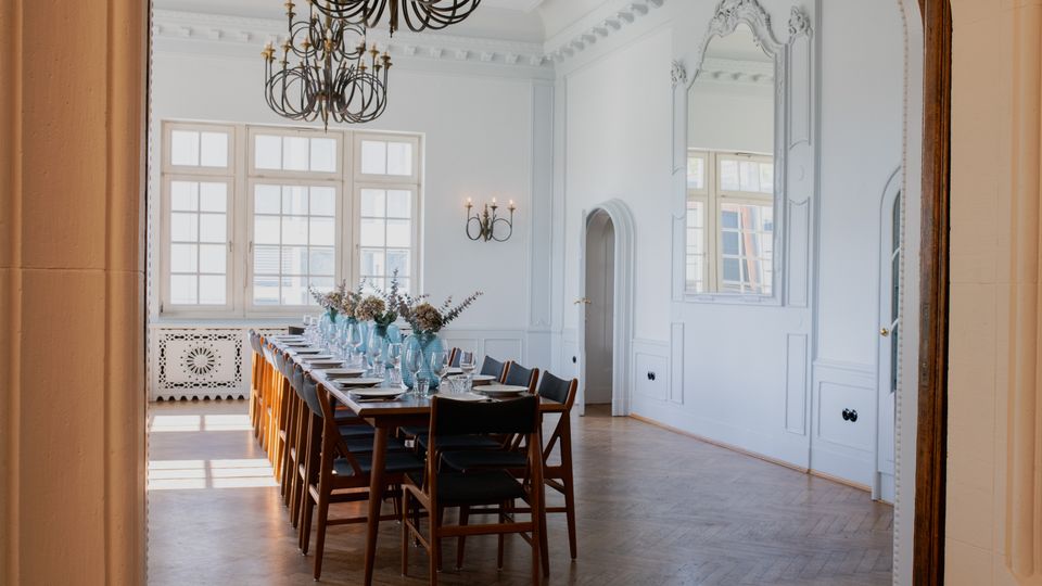 Eleganter Speisesaal mit einer langen gedeckten Tafel, dekoriert mit blauen Vasen und Trockenblumen. Der Raum ist lichtdurchflutet, hat Holzparkett, weiße Wände mit Stuckdetails, einen großen Spiegel sowie Kronleuchter und hohe Sprossenfenster.