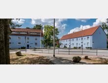 Wohngebiet mit zwei hellblauen, zweigeschossigen Gebäuden mit roten Satteldächern, umgeben von Straßen und Gehwegen. Im Vordergrund sind Bäume, Grünflächen und große Steine sichtbar, im Hintergrund Bäume und ein teils bewölkter Himmel.