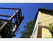Blick auf eine fragmentarische, rostige Metallkonstruktion links und eine verwitterte gelbe Hauswand mit Rissen rechts, vor einem klaren blauen Himmel und einem Baum im Hintergrund.