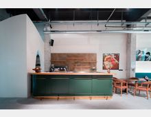 Moderne Café-Bar mit einer grünen Theke mit Holzdetails, dahinter eine Kaffeemaschine und Regale an einer Wand in Beton-Optik. Im Hintergrund befinden sich gerahmte Fotografien sowie ein Sitzbereich mit Polsterbänken und Holzmöbeln.