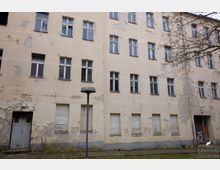 Fassade eines mehrstöckigen, verfallenen Wohngebäudes mit bröckelndem, beigem Putz, beschädigten Fenstern und teilweise heruntergelassenen Rollläden. Im Vordergrund sind eine Laterne, ein schmaler Gehweg und kahle Äste eines Baumes zu sehen.