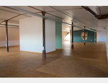 Großer, leerer Innenraum mit hellbraunem Holzboden, weißen und grünen Wänden sowie sichtbaren Metallstützen. An einer grünen Wand ist ein großes Wandbild einer stilisierten Hand erkennbar, die natürliche Beleuchtung kommt durch Fenster im Hintergrund.