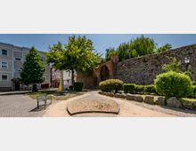 Ein kleiner öffentlicher Platz mit einem Sandspielbereich, einer Bank und Bäumen. Im Hintergrund ist ein historisches Tor aus rotem Backstein, eingebettet in eine Steinmauer, sowie benachbarte Wohngebäude zu sehen.