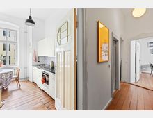 Helle Wohnung mit einem Flur aus Holzdielen, der in eine Küche und ein angrenzendes Zimmer führt. Die Küche hat weiße Schränke, eine Arbeitsplatte mit Spüle und Herd, ein Fenster sowie einen Holztisch mit Stühlen; an der Wand im Flur hängt ein auffälliges, orange gerahmtes Bild.