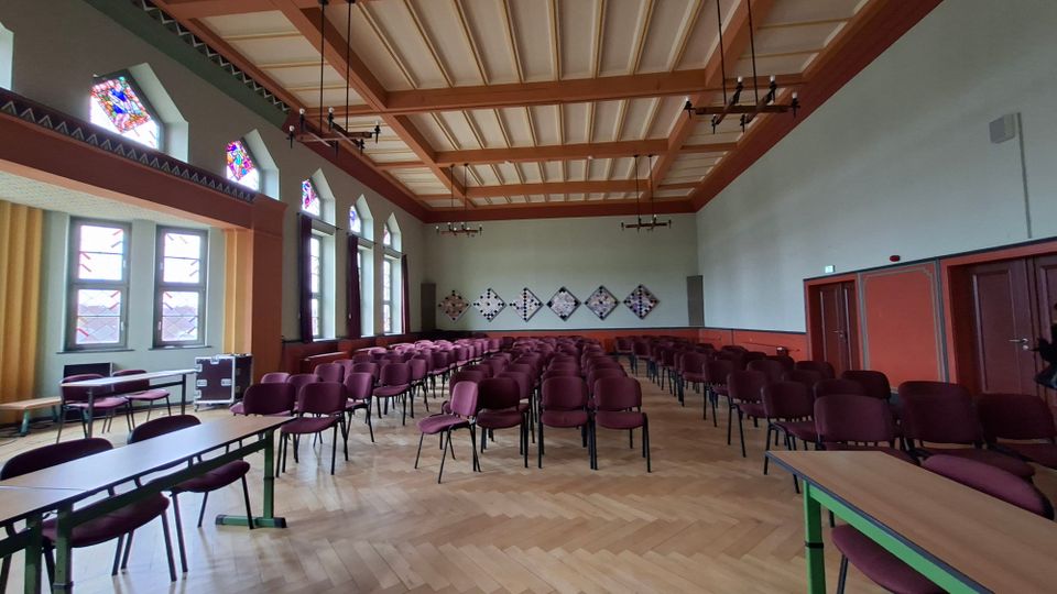 Großer Veranstaltungs- oder Sitzungssaal mit Reihen aus bordeauxroten Stühlen auf hellem Fischgrätparkett und einem hohen Kassettendeckel aus Holz. An der linken Seite stehen hohe Fenster mit bunten Glasoberlichtern, an der Stirnwand hängen rautenförmige Wandtafeln.