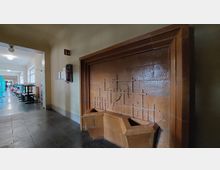 Ein Innenraum mit einem braunen Wandbrunnen aus Keramik mit geometrischen Mustern. Im Hintergrund ein Flur mit Tischen, Stühlen und blauen Trennwänden sowie Fenstern, die Tageslicht einlassen.