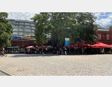 Offener Platz mit gepflastertem Boden vor einem historischen Backsteingebäude, das teilweise von Bäumen verdeckt ist. Im Vordergrund befinden sich ein Fahrradfahrer, Café-Tische mit Sonnenschirmen sowie ein Schild mit Informationen, im Hintergrund fährt eine S-Bahn auf einer erhöhten Trasse.