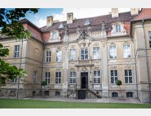 Historische Fassade eines barocken Gebäudes mit symmetrischer Architektur, verzierten Fensterrahmen, Stuckornamenten und Figuren auf dem Dach. Im Vordergrund befindet sich ein Rasen mit vereinzelten Bäumen.