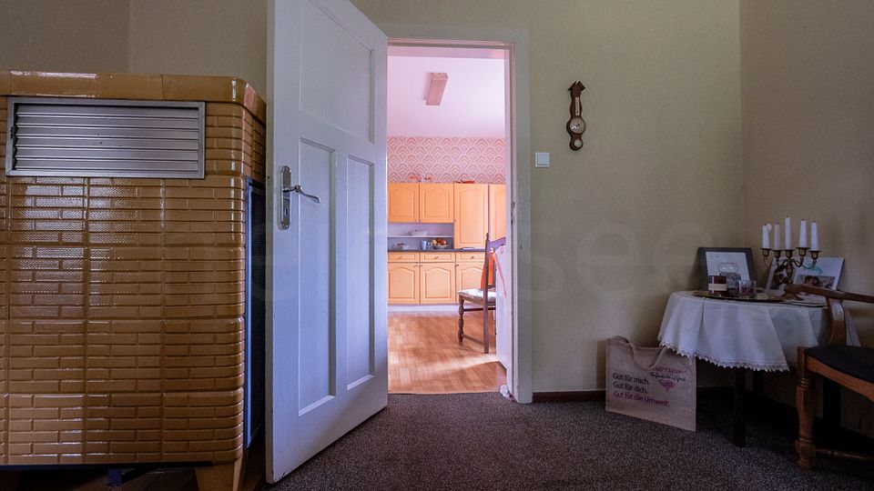 Blick aus einem Wohnraum mit braun gefliestem Kachelofen und kleinem Tisch mit Kerzenständer durch eine offene weiße Tür in eine helle Küche. In der Küche sind helle Holzschränke, gemusterte Tapete und ein Holzboden zu sehen.