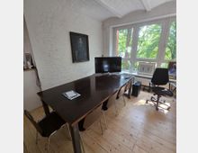 Ein kleiner Besprechungsraum mit einem großen dunklen Holztisch und mehreren Stühlen, umgeben von weiß gestrichenen Backsteinwänden. Ein großes Fenster mit Blick auf grüne Bäume bringt viel Licht in den Raum; an den Wänden hängen Bilder, und auf dem Tisch steht ein Monitor.