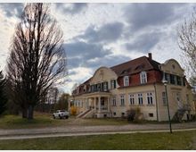Großes, zweigeschossiges Herrenhaus mit hellem Putz, rotem Mansarddach und mehreren Gaubenfenstern, davor eine Treppe zu einem überdachten Eingang. Das Gebäude steht in einer offenen Grünanlage mit hohen, unbelaubten Bäumen, einem Kiesweg und bewölktem Himmel.