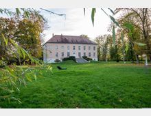 Helles Herrenhaus mit rosa Fassade und roten Dachziegeln, umgeben von einem weitläufigen, gepflegten Rasen und Herbstbäumen; eine Treppe führt zum Haupteingang der symmetrischen Fassade.