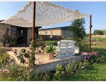 Eine Terrasse mit Backsteinboden und Metallstützen, überdacht von einem weißen Netz-Sonnensegel. Im Vordergrund stehen Pflanzen, Blumenbeete und eine Sitzbank aus weißen Holzpaletten. Im Hintergrund ist ein Haus aus hellen Ziegeln und ein Garten mit Rasen und Teich erkennbar.