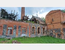 Ruine eines roten Ziegelgebäudes mit zerfallenen Dächern und einem hohen Schornstein. Die Fassaden bestehen aus einer Kombination aus Backstein und Naturstein, mit teils verfallenen Fenstern und einer überwucherten Umgebung.