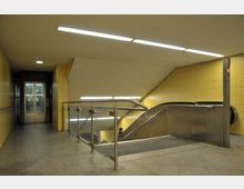 Untergrundstation mit gelb gekachelten Wänden, einer Rolltreppe nach unten und einem Aufzug auf der linken Seite. Der Boden ist mit grauem Steinbelag ausgelegt, und Metallgeländer trennen die Bereiche voneinander.