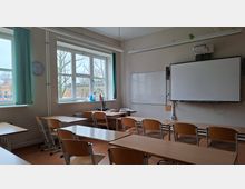 Leerer Klassenraum mit Reihen aus Holztischen und -stühlen, großen Fenstern mit grünen Vorhängen und einer Wanduhr. Vorn befinden sich eine Tafel bzw. ein interaktives Whiteboard mit Deckenprojektor sowie ein Lehrerpult.