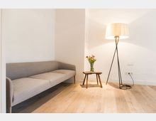 Ein minimalistisch eingerichteter Raum mit heller Holzoptik, einer grauen Polstercouch, einem kleinen hölzernen Beistelltisch mit Blumenstrauß und einer hohen Stehleuchte mit schlichten Metallbeinen und einem hellen Lampenschirm. Die Wände sind weiß und schlicht gehalten.