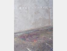 Eine raue Betonwand mit Spuren von Verfärbungen und Ausbesserungen sowie ein fleckiger Betonboden, der mit bunten Farbspritzern und -flecken bedeckt ist. Die Szene könnte ein Atelier oder eine Werkstatt darstellen.