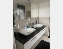 Modernes Badezimmer mit einem Doppelwaschbecken auf einer schwarzen Arbeitsplatte, darunter weiße Schubladen. Im Hintergrund befinden sich ein großer Spiegel, hellgraue Wandfliesen und ein Fenster mit geschlossenen Jalousien.