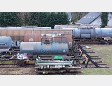 Güterwagen-Anlage mit mehreren abgestellten Kesselwagen auf parallel verlaufenden Gleisen, umgeben von Bäumen und einigen Wohnhäusern im Hintergrund. Die Kesselwagen sind teilweise mit Graffiti versehen und zeigen Gebrauchsspuren.