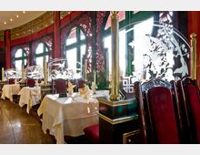 Restaurant mit asiatischem Design, geprägt von dunkelroten Stühlen, weißen Tischdecken und kunstvoll geschnitzten Holzelementen an den runden Fenstern. Dekorative Glastrennwände mit floralen Gravuren und goldenen Säulen trennen die Tische voneinander.