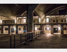 Industriehalle mit gewölbter Decke, mehreren Ebenen und kargen Betonwänden. Der Boden ist gepflastert, und der Raum wird durch gelbe Beleuchtung und eine Reihe von Kerzen entlang eines Metallgeländers erhellt.