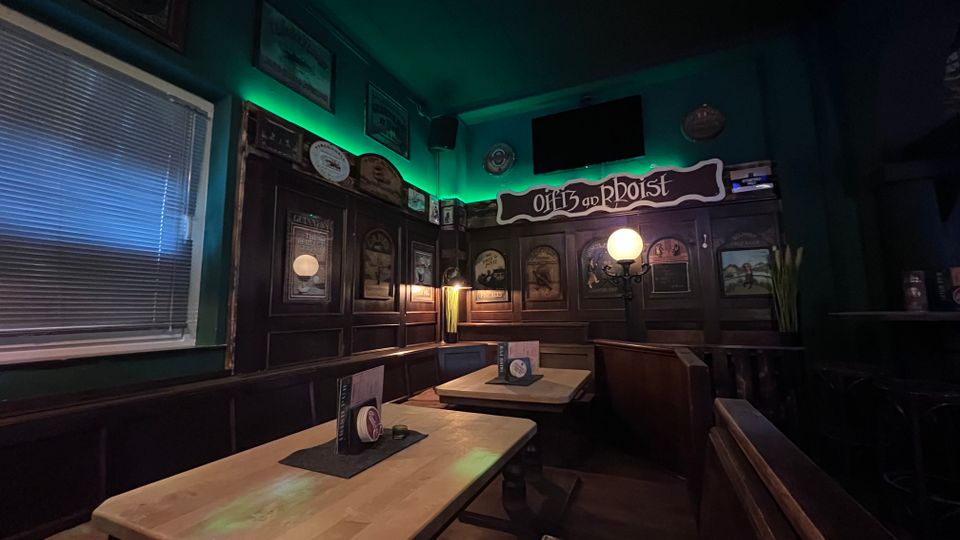 Gemütlicher Pub-Innenraum mit dunkler Holzvertäfelung, Eckbänken und mehreren Holztischen, auf denen Speisekarten stehen. An den Wänden hängen gerahmte Bilder und ein Schild „Off to an Rboist“, dazu gedämpfte Lampen und grüne indirekte Beleuchtung unter der Decke.