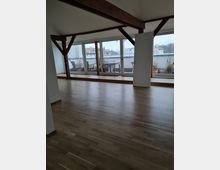 Ein leerer, lichtdurchfluteter Raum mit Holzfußboden, sichtbaren Holzträgern und einer großen Fensterfront, die auf eine Terrasse mit Pflanzen und eine städtische Umgebung im Hintergrund blickt.