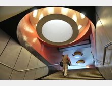 Treppe in einer U-Bahn-Station mit auffälliger, kreisförmiger Deckenbeleuchtung und roten Mosaikfliesen an den Wänden. Ein Mann steigt die Treppe hinauf in Richtung eines hell erleuchteten Bereichs.