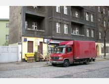 Wohnhaus mit dunkelgrauer Fassade und weißen Fenstern, im Erdgeschoss befinden sich ein Laden für Spirituosen sowie geschlossene Rollläden. Vor dem Gebäude steht ein roter Lieferwagen auf einer gepflasterten Straßenfläche, daneben gelbe Mülltonnen und gestapeltes Holz.