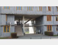 Mehrstöckiges, modernes Gebäude mit grauer Fassade und gelb umrandeten Fenstern. In der Mitte des Gebäudes befindet sich ein überdachter Durchgang mit einer Treppe, die zu einem offenen Bereich im Obergeschoss führt; im Hintergrund sind Bäume sichtbar.