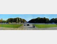 Weitläufige Straßenkreuzung mit Bäumen im Hintergrund, Rasenflächen im Vordergrund und einer gepflasterten Fläche mit Treppenzugang in der Mitte; mehrere Flaggenmasten stehen links der Szene, und Fahrzeuge sind auf den Straßen zu sehen.